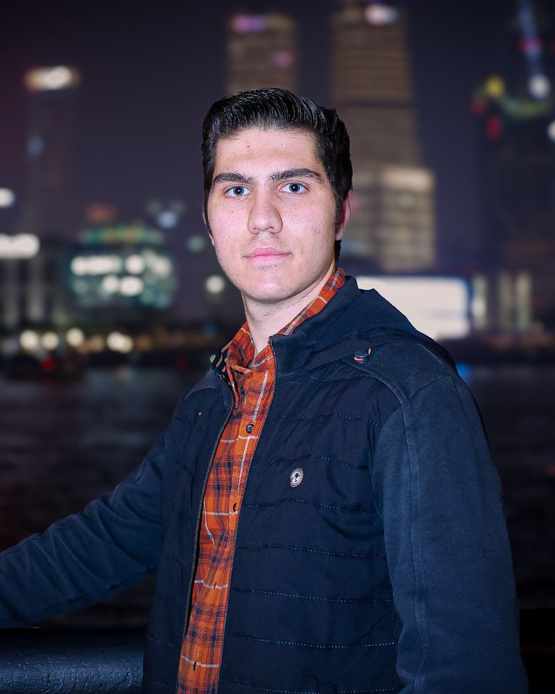 Ivan Maggini di notte, in giacca blu e camicia a quadri arancione, con le luci di una città sfocate alle spalle.