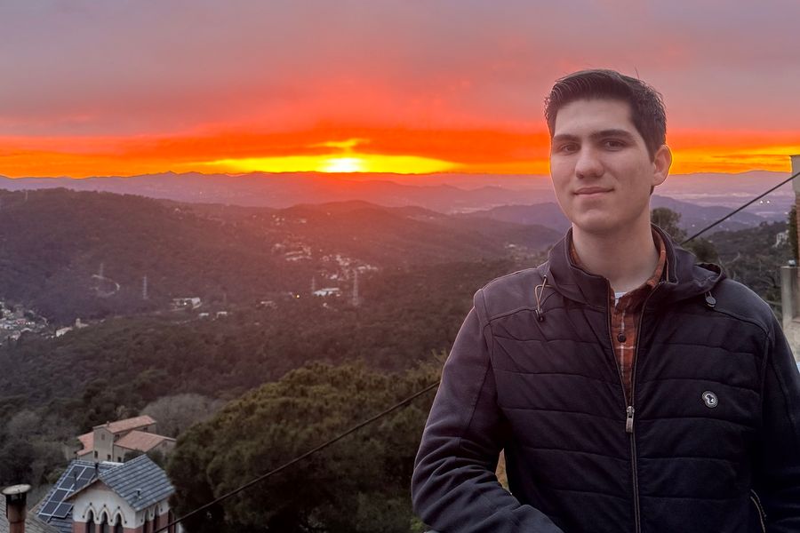 Ivan Maggini sorride appoggiato a una ringhiera, con un tramonto arancione sulle colline alle spalle.